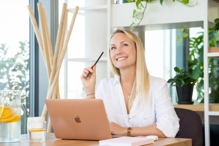 Eine Frau sitzt lächelnd mit einem Stift in der Hand am Laptop und schaut in die Kamera