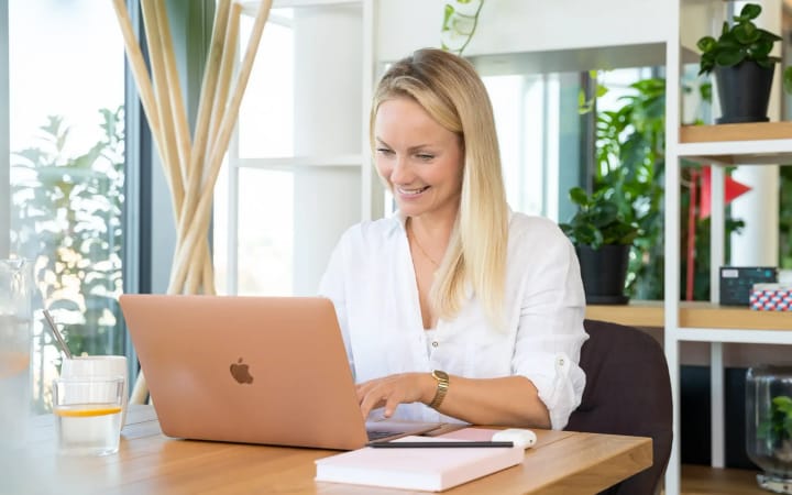 Eine Frau tippt konzentriert am Laptop, daneben liegt ein Notizbuch auf einem Holztisch