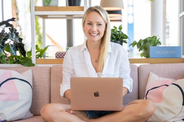 Eine Frau sitzt im Schneidersitz auf einem rosa Sofa und arbeitet lächelnd am Laptop