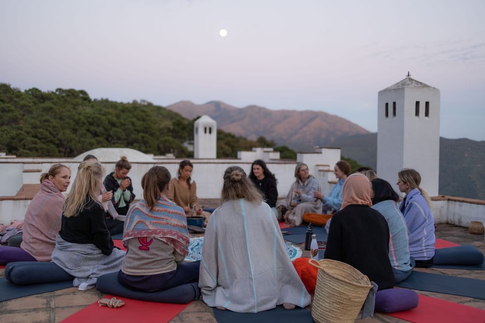 Teilnehmerinnen sitzen in der Abenddämmerung im Kreis auf einer Dachterrasse, im Hintergrund Hügel und ein weißer Turm