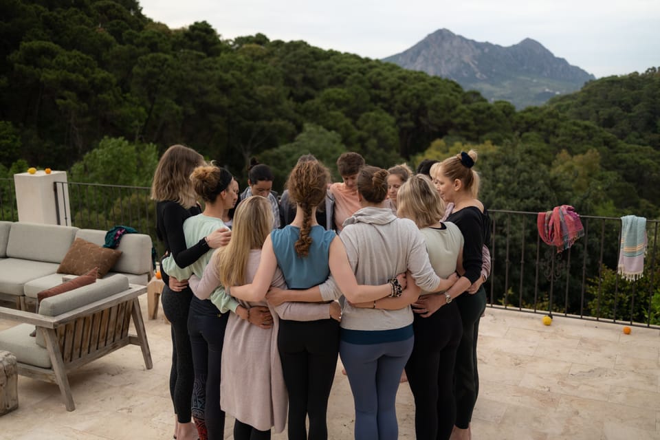Eine Gruppe steht Arm in Arm im Kreis auf einer Terrasse, von hinten fotografiert, dahinter Wald und Berge