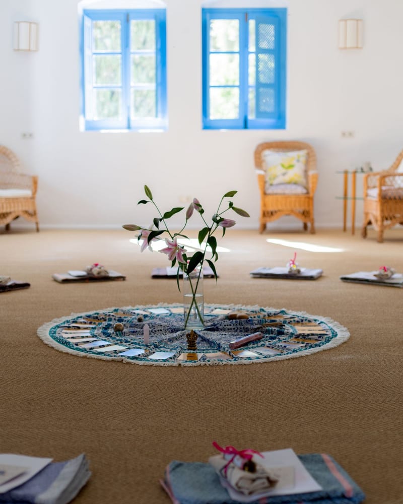 Heller Yogaraum mit blauen Fenstern, in der Mitte eine Mandala-Decke mit Lilien, rundherum Kissen und Decken